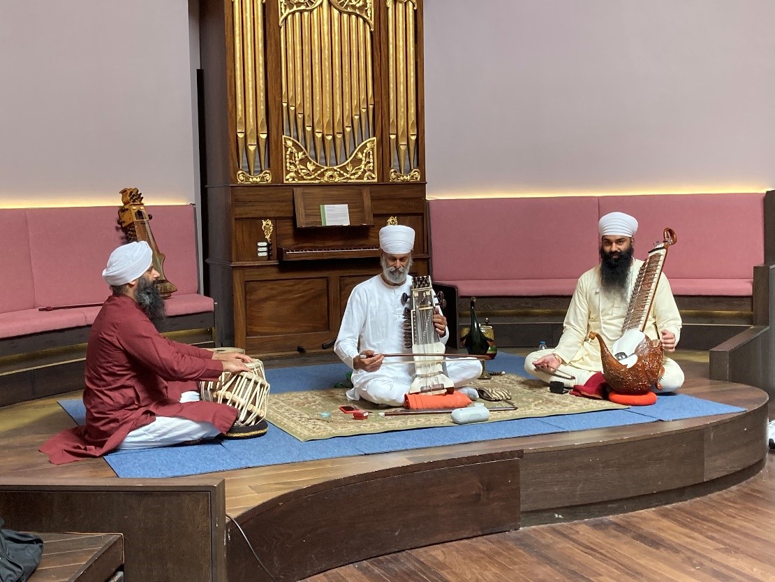 Figure 1 Sodhi Deerhe, Surjit Singh, and Avtar Singh play the tabla, sarangi, and taus Three men are sitting on the concert room stage playing instruments from India. The men are Sodhi Deerhe, Surjit Singh, and Avtar Singh. They are playing the tabla, sarangi, and taus.